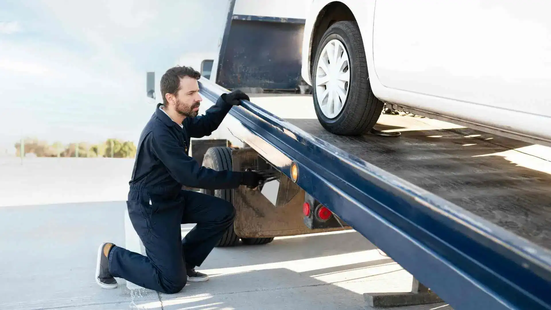 Tow truck worker kneels on a flatbed ramp, guiding a white car onto the truck bed while securing it with gloves on.