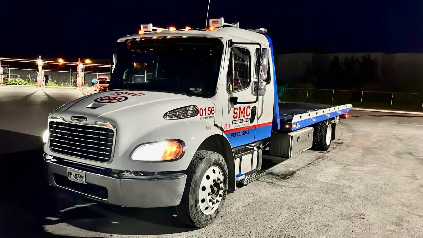 Tow truck with blue flatbed parked at night, SMC Towing Group logo on the door and roof lights.