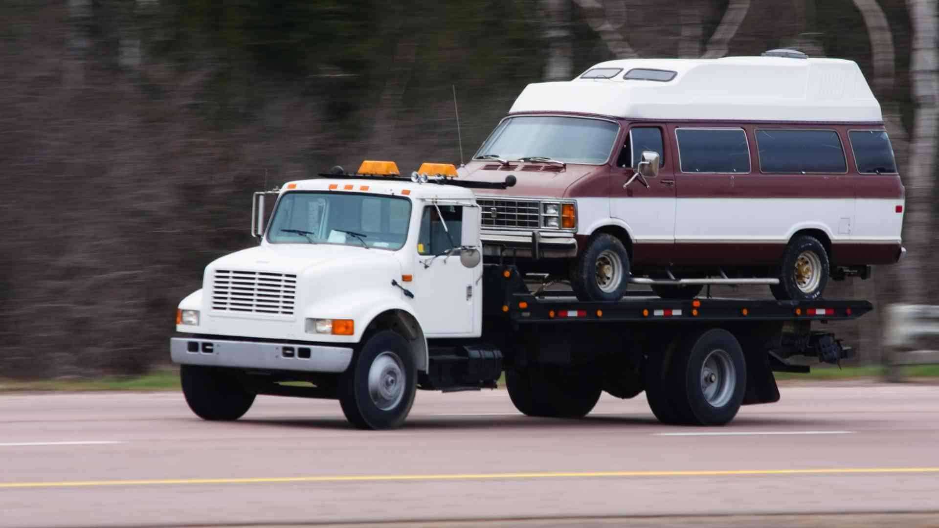 Tow truck hauling a two-tone white and burgundy passenger van on a flatbed trailer, moving down a road.