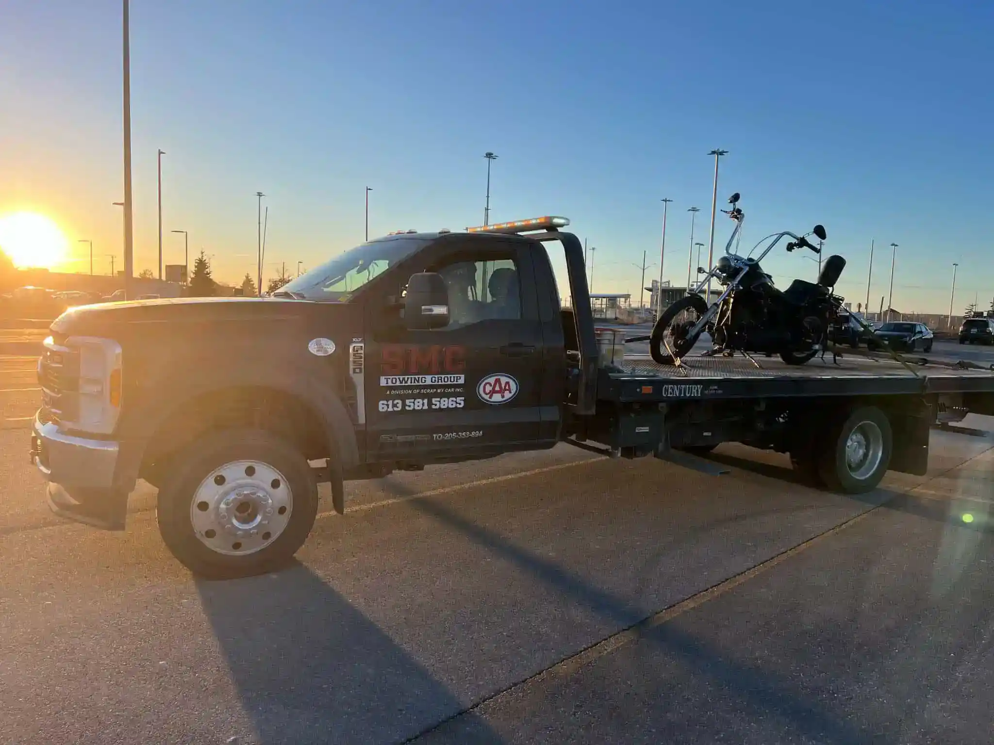 Tow truck with a motorcycle secured on a flatbed in a parking lot at sunset, with bright sun on the left and streetlights in the background.