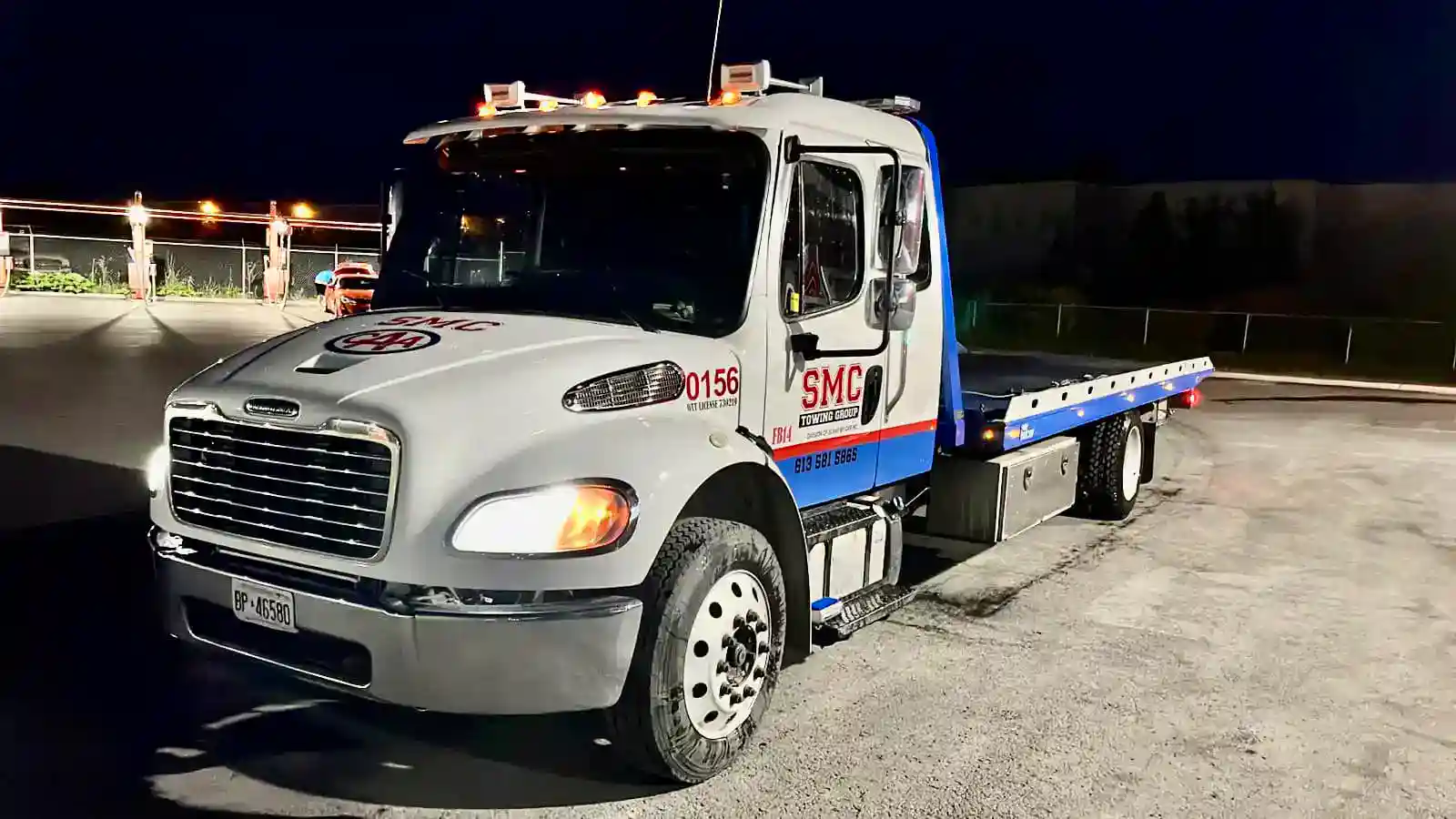 White tow truck with a blue flatbed parked at night; headlights on and emergency lights on top, labeled 'SMC Towing Group' on the side, ready to tow a vehicle.
