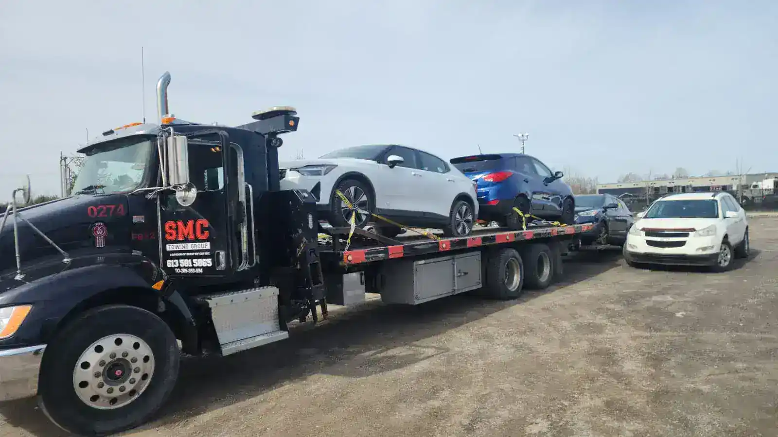 Black tow truck from SMC Towing Group on a flatbed trailer carrying three cars—a white sedan, a blue hatchback, and a gray car—on a dirt lot.