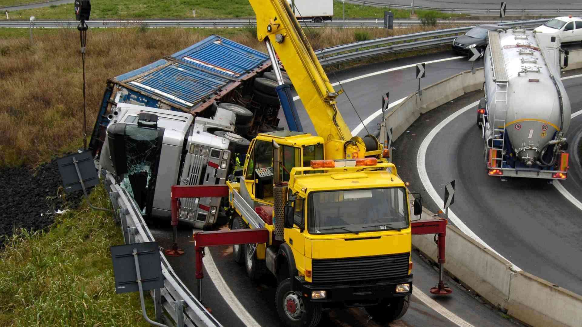 Crane tow truck lifting an overturned truck on a curved highway ramp beside a tanker trailer and barriers, with debris around.