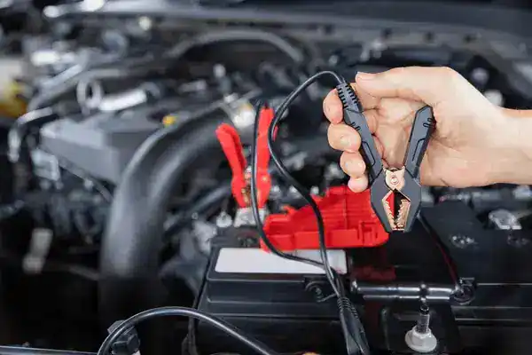 Person holds jumper cables with red and black clamps near a car battery under the hood, preparing to jump-start the vehicle.